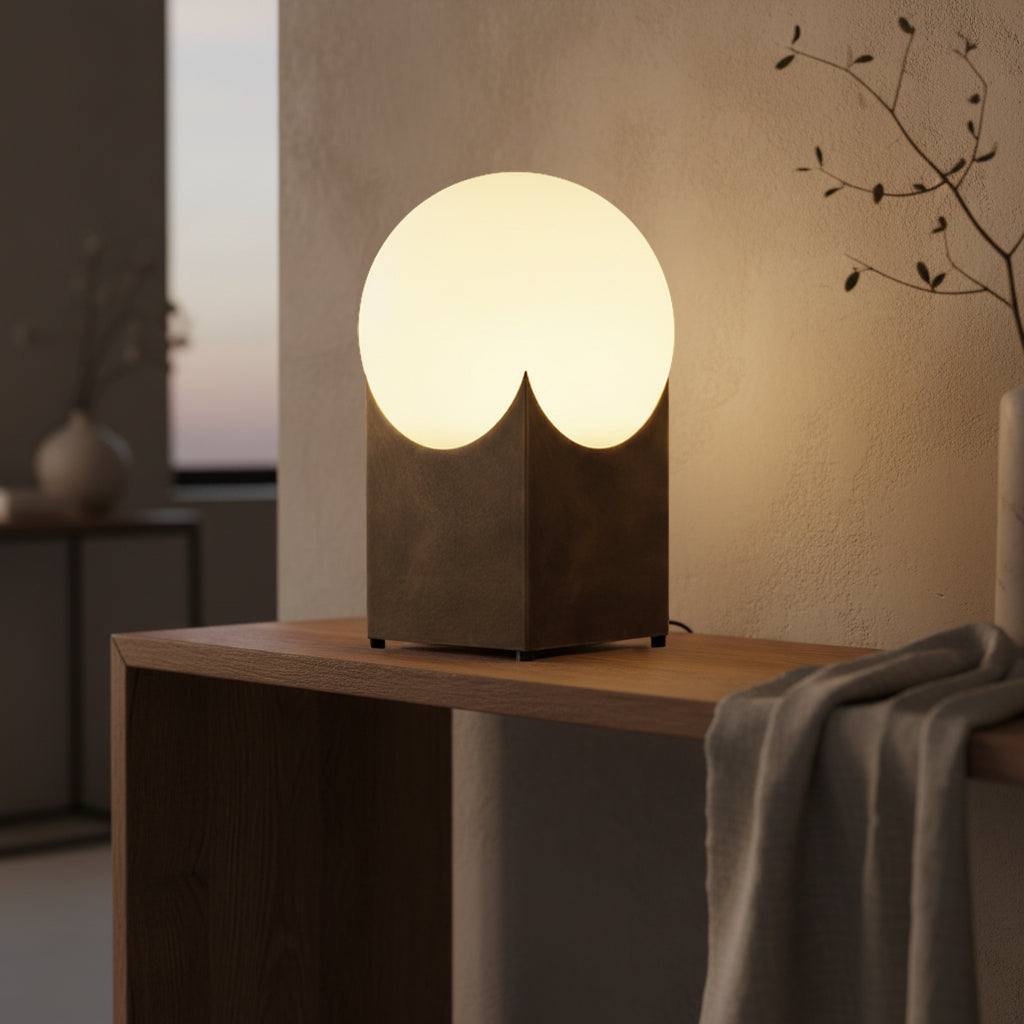 A modern table lamp with a glowing round frosted shade and a dark textured geometric base, placed on a wooden table in a softly lit interior room.
