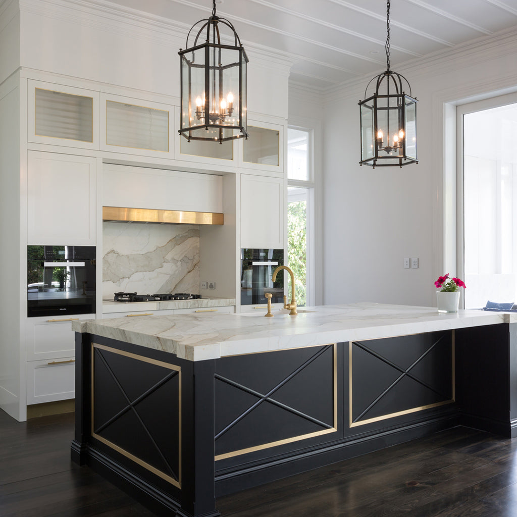 A modern kitchen island with a black base, gold trim, and a white marble countertop, set in a bright kitchen with white cabinets, dark wood floors, and two black pendant lights.
