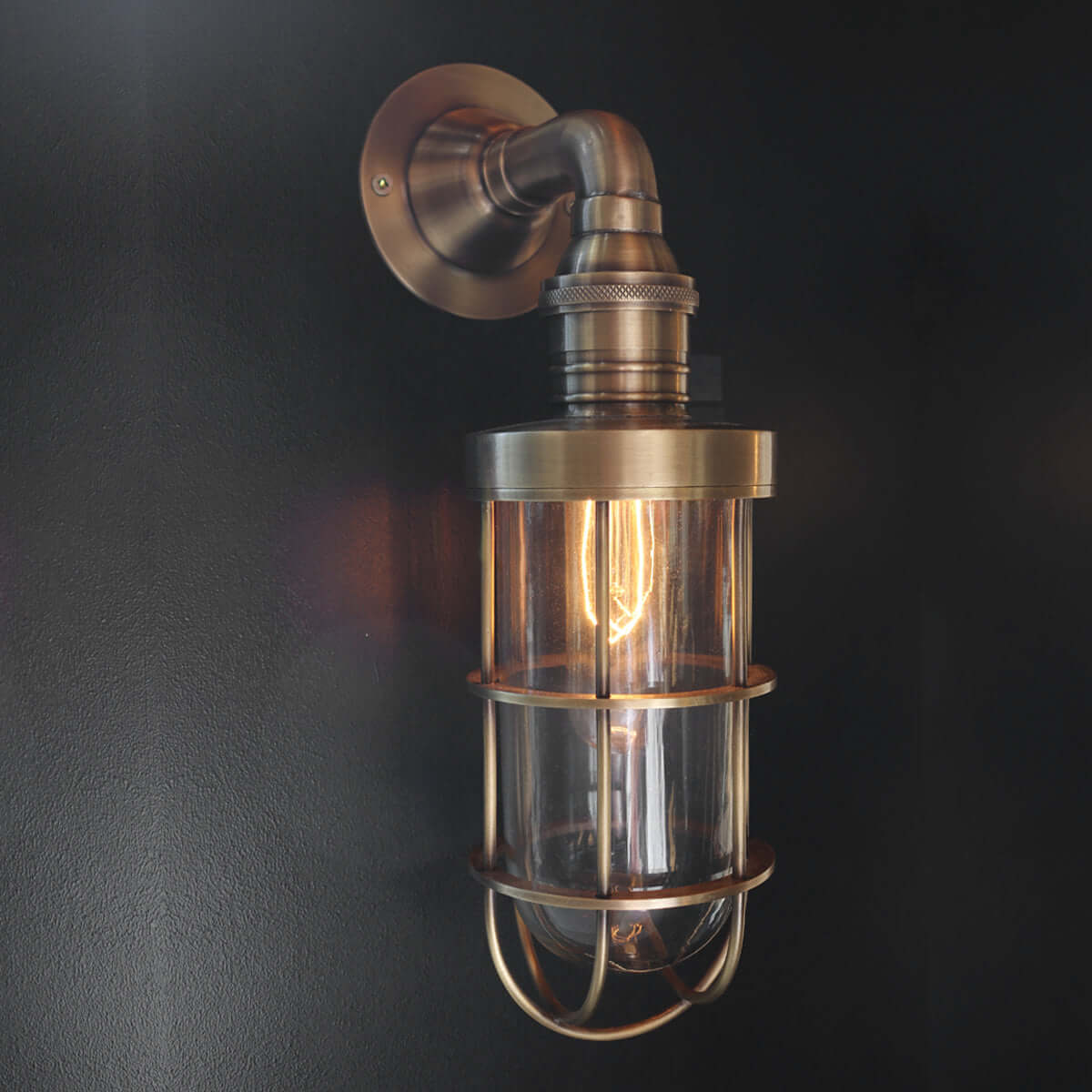Industrial-style wall sconce with a brushed brass metal mount, clear glass shade, and visible warm Edison bulb against a dark wall.