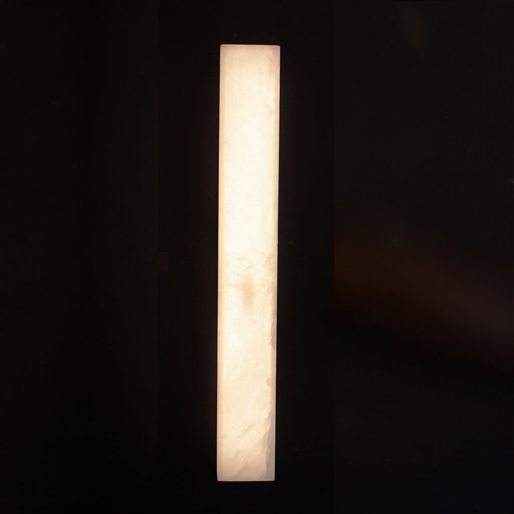 A tall, narrow rectangular wall sconce made of glowing white stone, emitting a warm light against a very dark background.
