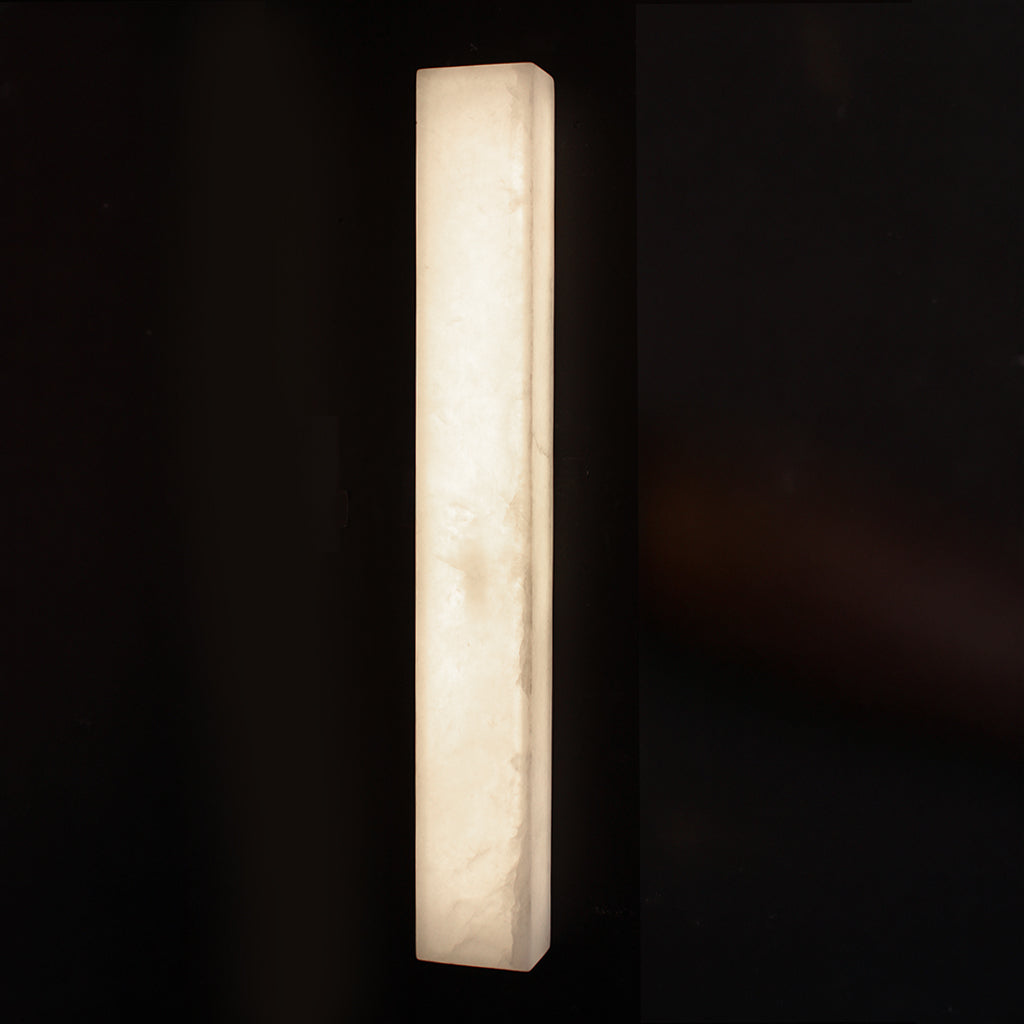 A modern rectangular wall sconce made of glowing warm white stone, shown against a very dark black background.