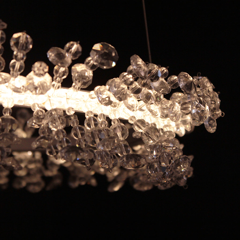 A modern circular LED chandelier with clear crystal beading, suspended from a silver ceiling canopy against a dark background.
