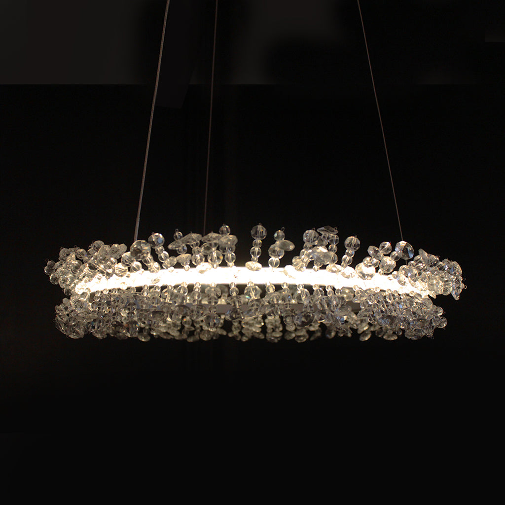 A modern horizontal crystal chandelier with a glowing white LED light ring and clear faceted crystal beads, suspended against a solid black background.