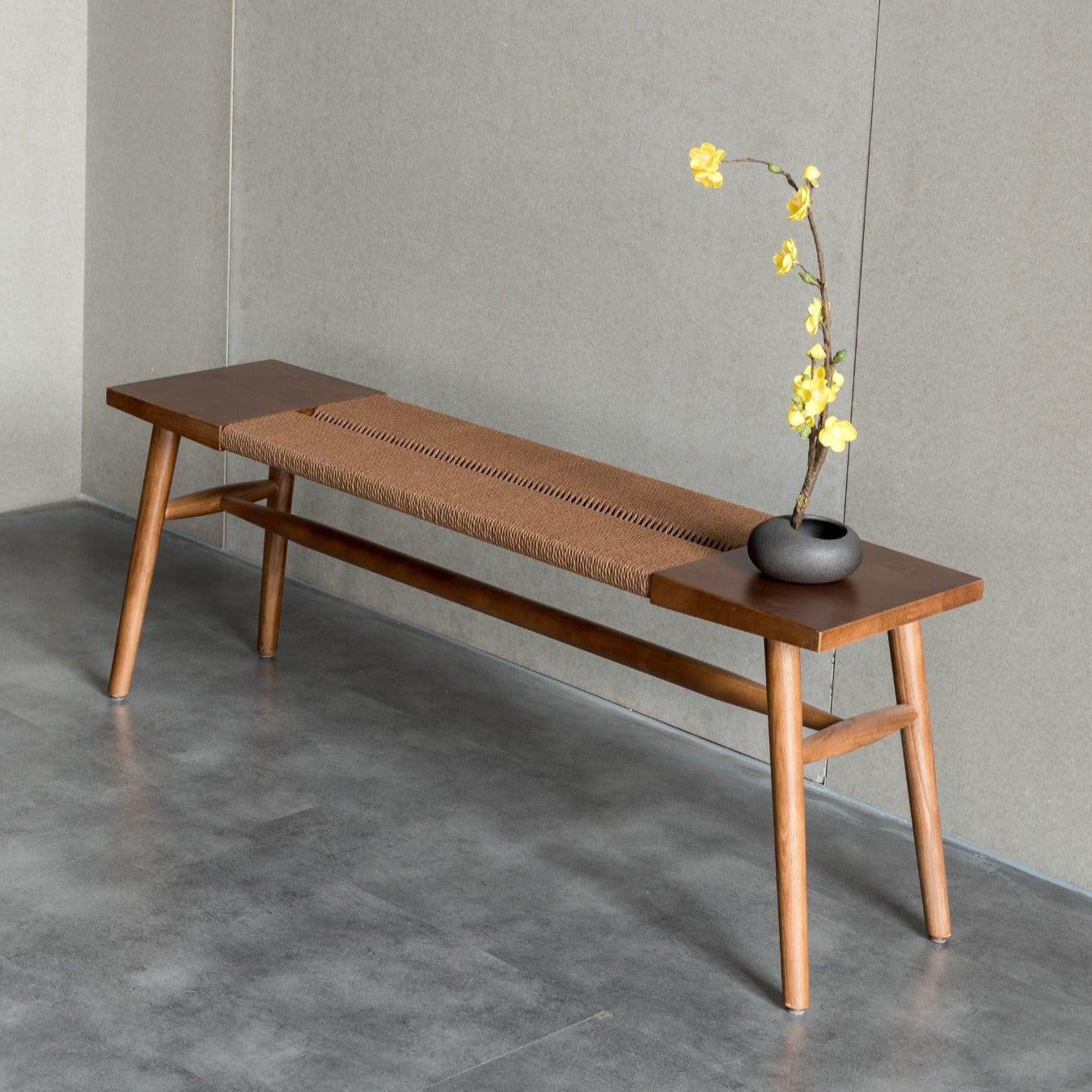 A Japandi Rope Woven Dining Bench with a walnut finish and handwoven rope centre, placed against a wall with a vase of yellow flowers on top.