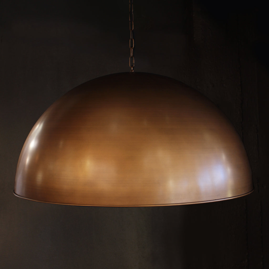 A large dome-shaped pendant light in a brushed brass or bronze finish, suspended by a chain against a dark, textured background.
