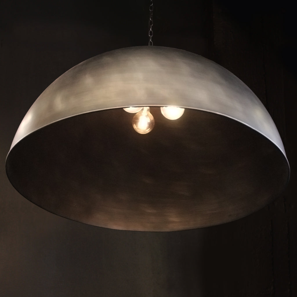 A large dome-shaped pendant light with a brushed silver metallic finish, shown illuminated against a dark background.