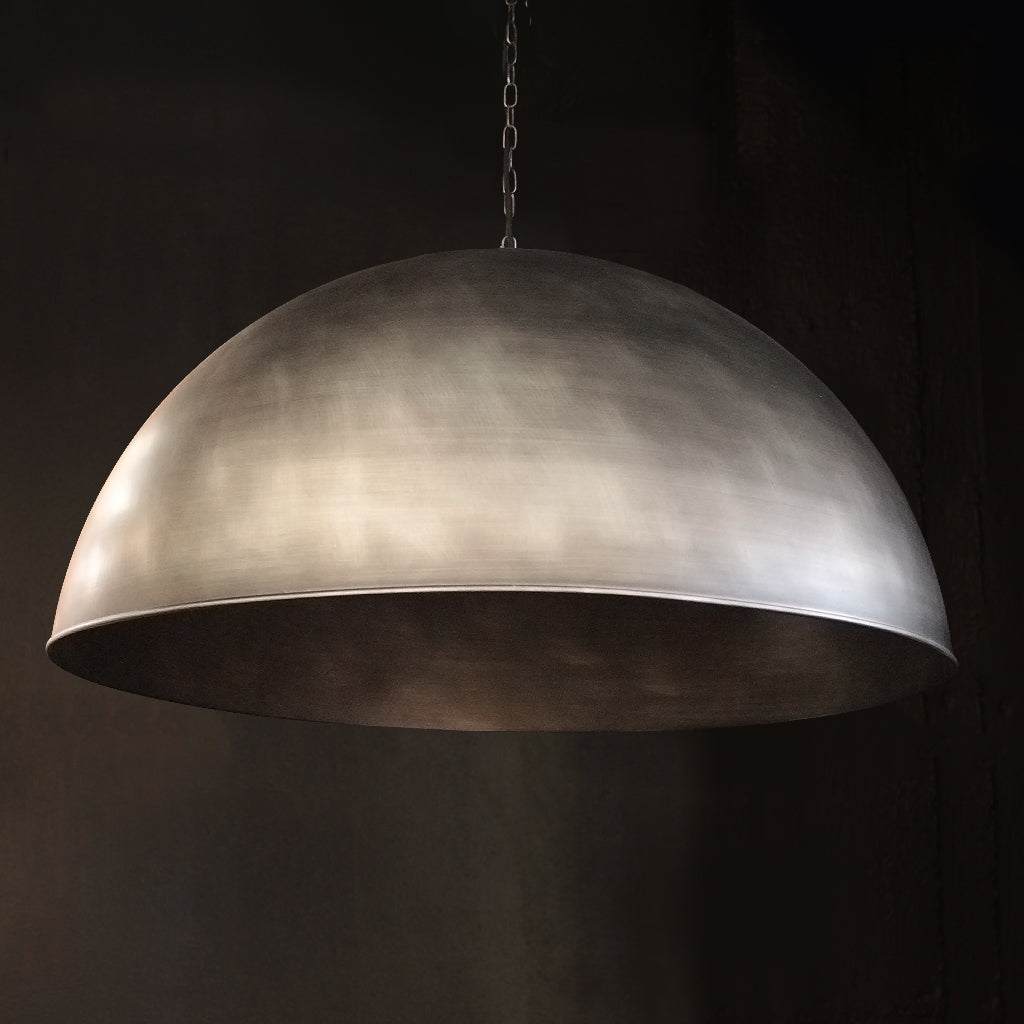 A large dome-shaped pendant light with a brushed metallic silver finish, suspended by a chain against a dark background.
