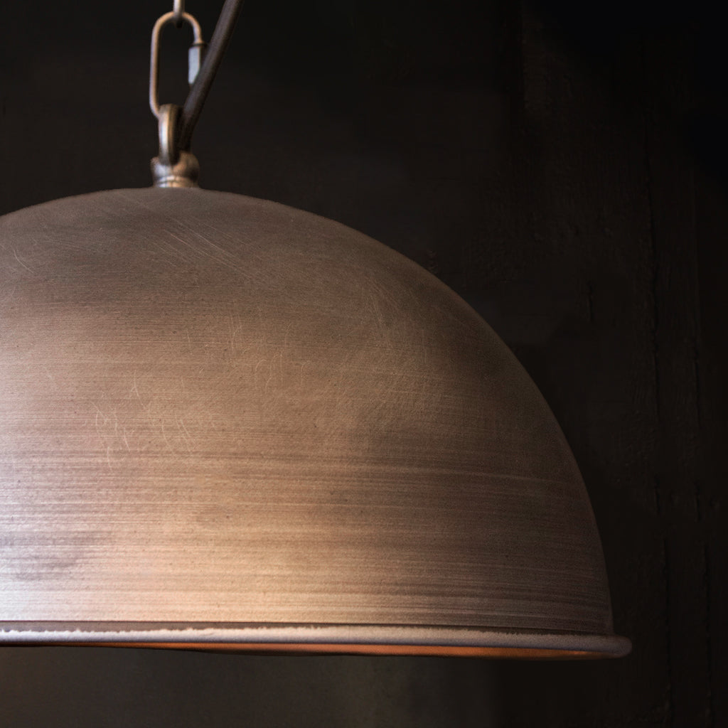 A close-up of a dome-shaped pendant light with a distressed nickel metal finish, suspended by a metal chain against a dark background.