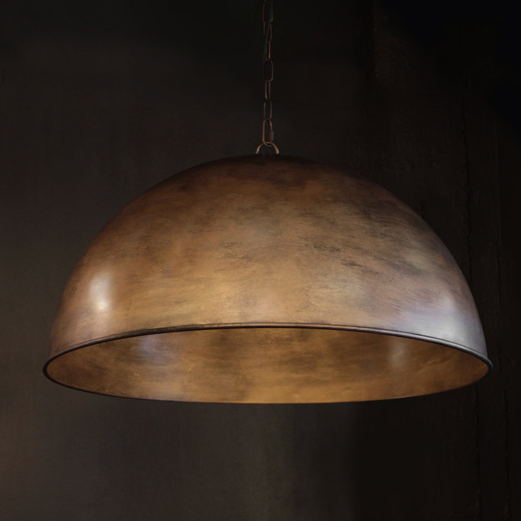 A large dome-shaped pendant light with a textured metallic brass finish, suspended by a thin chain against a dark, moody background.