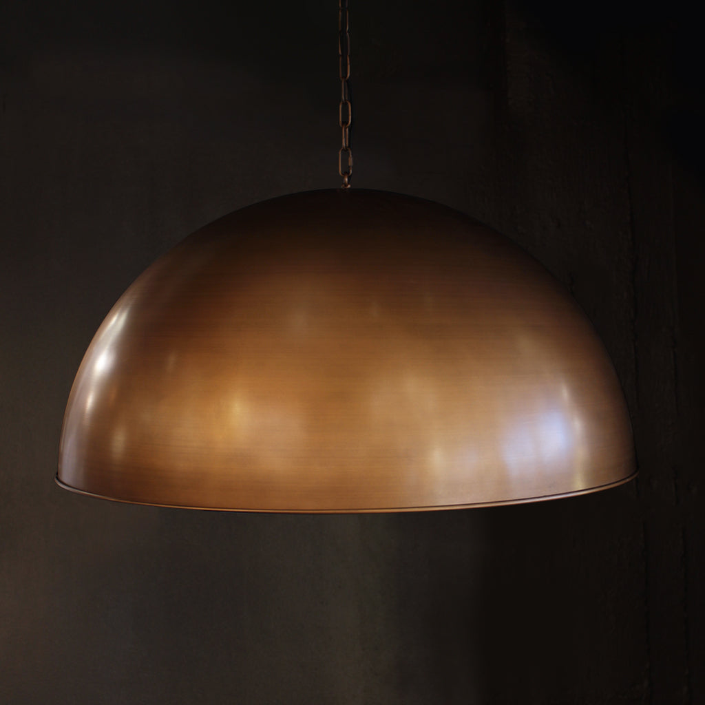 A large dome-shaped pendant light with a brushed antiqued brass finish, suspended against a dark, textured background.