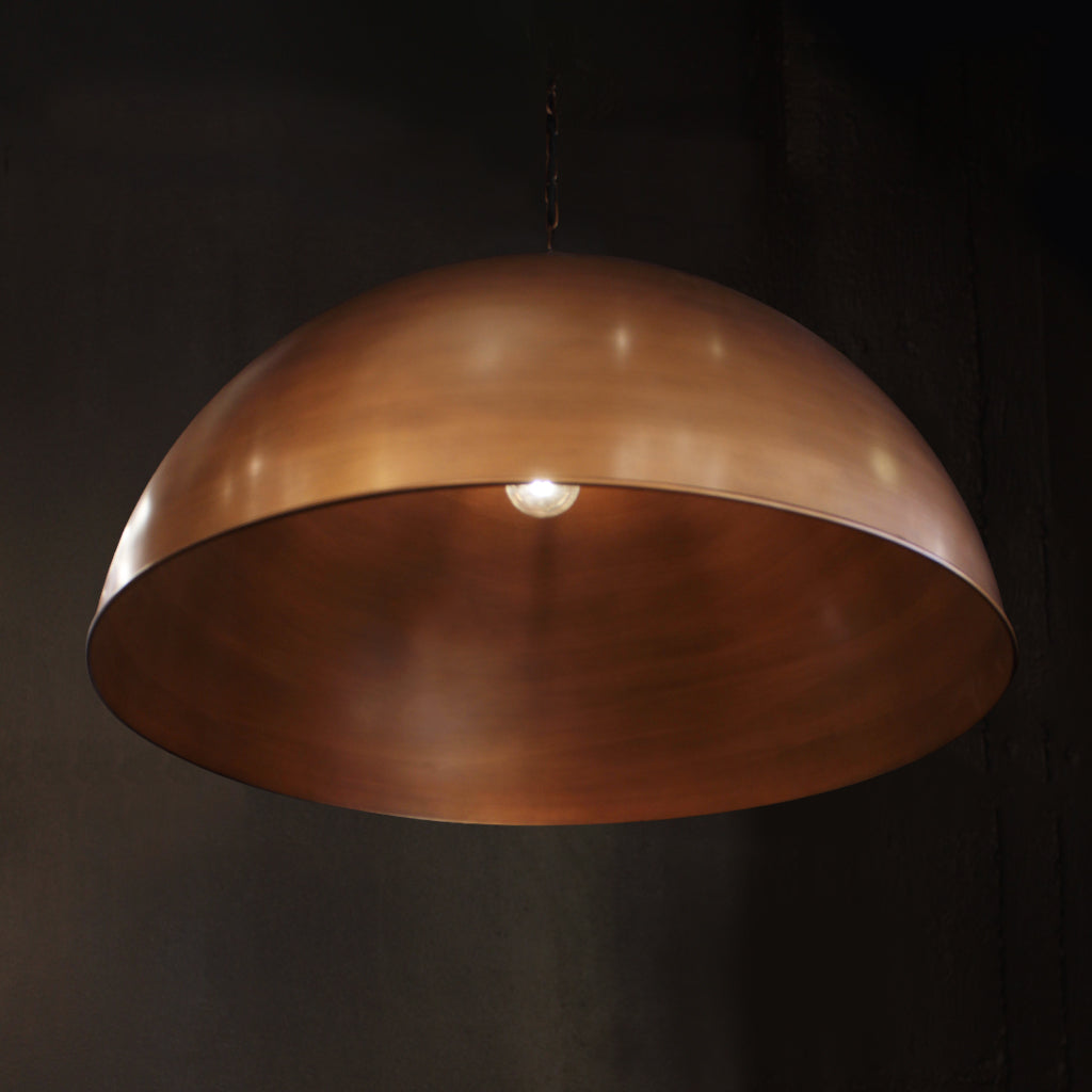 A large dome-shaped pendant light with a brushed antiqued brass finish, suspended against a dark, textured background.