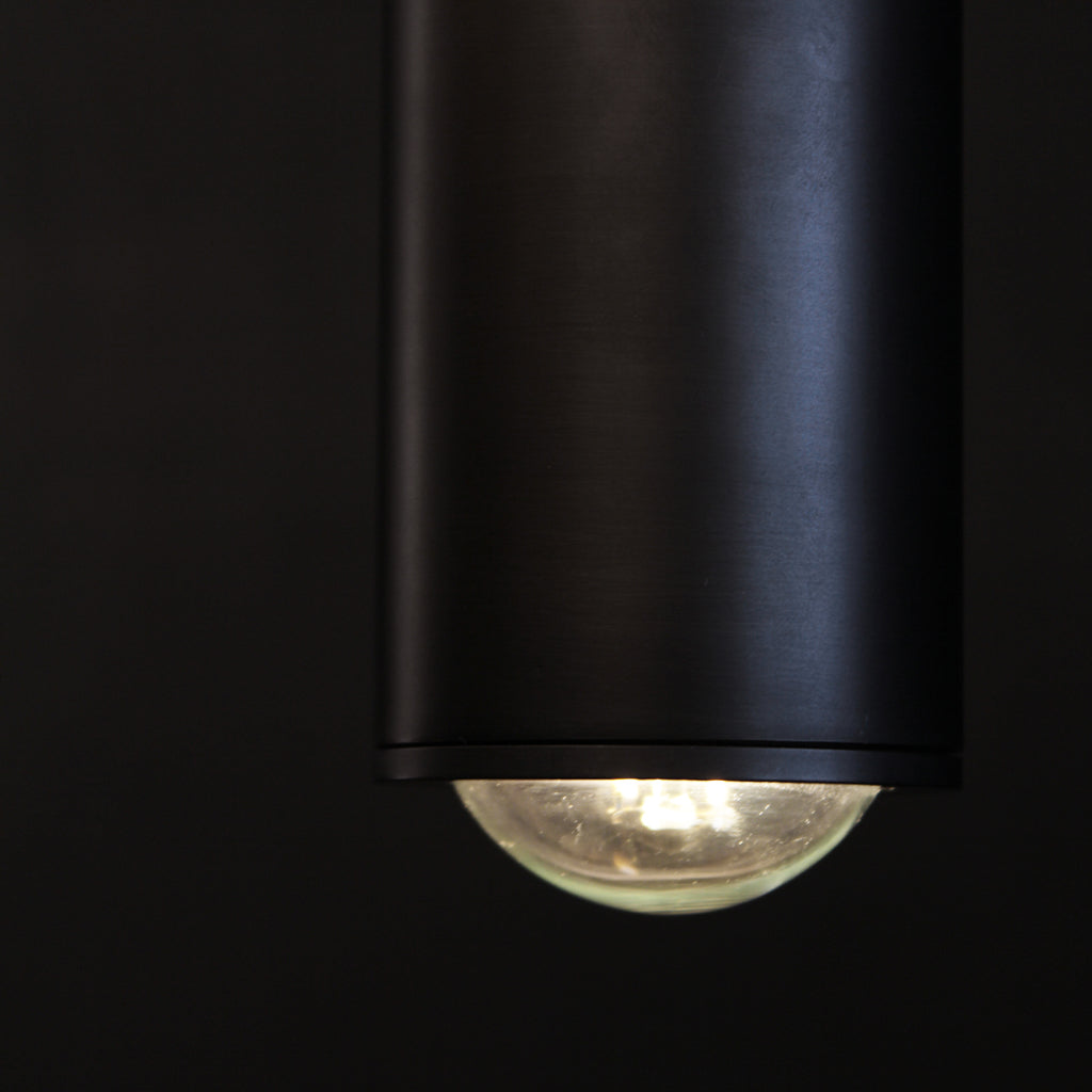 A modern gunmetal dark cylindrical pendant light hanging in front of a dark background