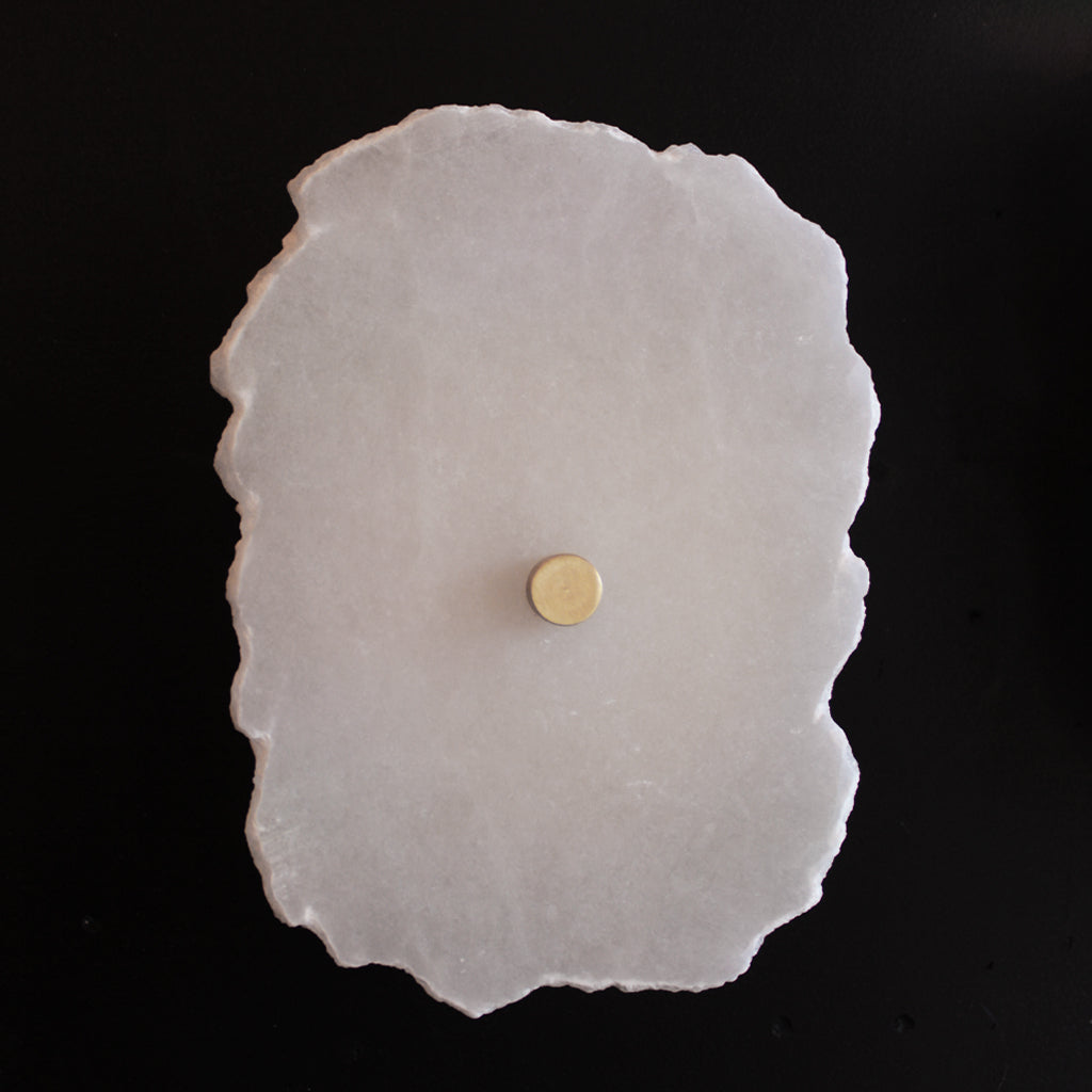 A raw, irregularly shaped white stone wall light with natural edges, featuring central brass detail, set against a solid black background.
