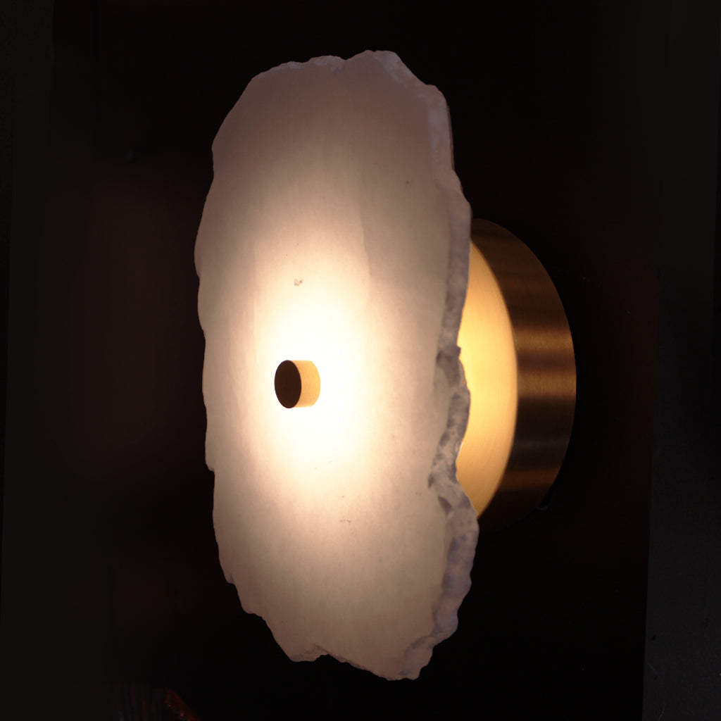 A modern wall sconce featuring a glowing, irregularly shaped white alabaster stone with a brushed brass metal backing and a central brass circular detail, set against a dark background.