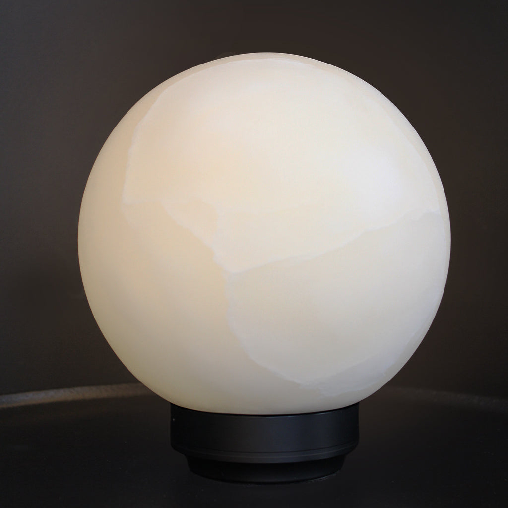 A spherical white alabaster lamp shade glowing with a warm light, mounted on a black circular base against a dark background.