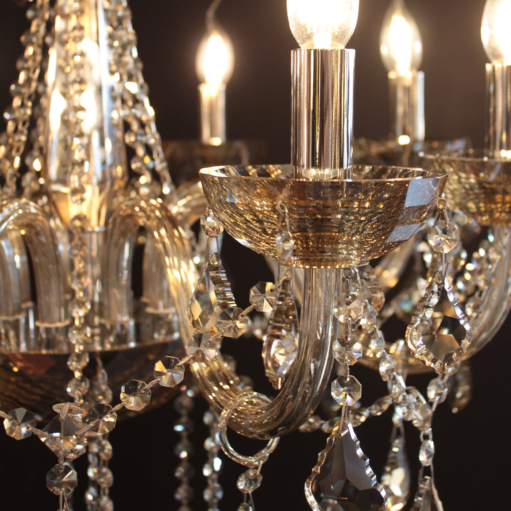 Close-up of a chandelier with amber glass crystal drops, curved amber glass arms, and gold-toned bowls, shown lit against a dark background.