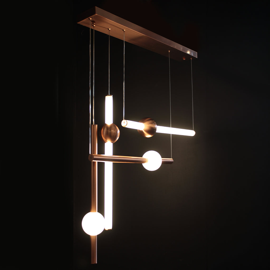 A modern geometric pendant light fixture with a brushed copper finish, featuring illuminated cylindrical tubes, spherical bulbs, and angled bars suspended against a solid black background.