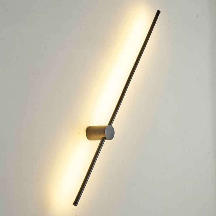 A modern linear wall sconce with a brushed brass cylindrical center and a long black rod, emitting a warm yellow light against a plain off-white wall.