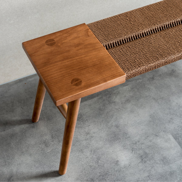 A Japandi Rope Woven Dining Bench with a walnut finish and handwoven rope centre, placed against a wall with a vase of yellow flowers on top.
