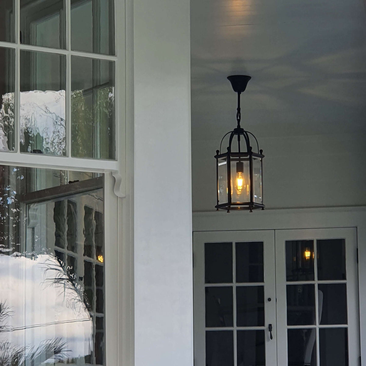 A black metal lantern-style pendant light with clear glass panels hangs from a ceiling on a porch with white-framed windows and doors.